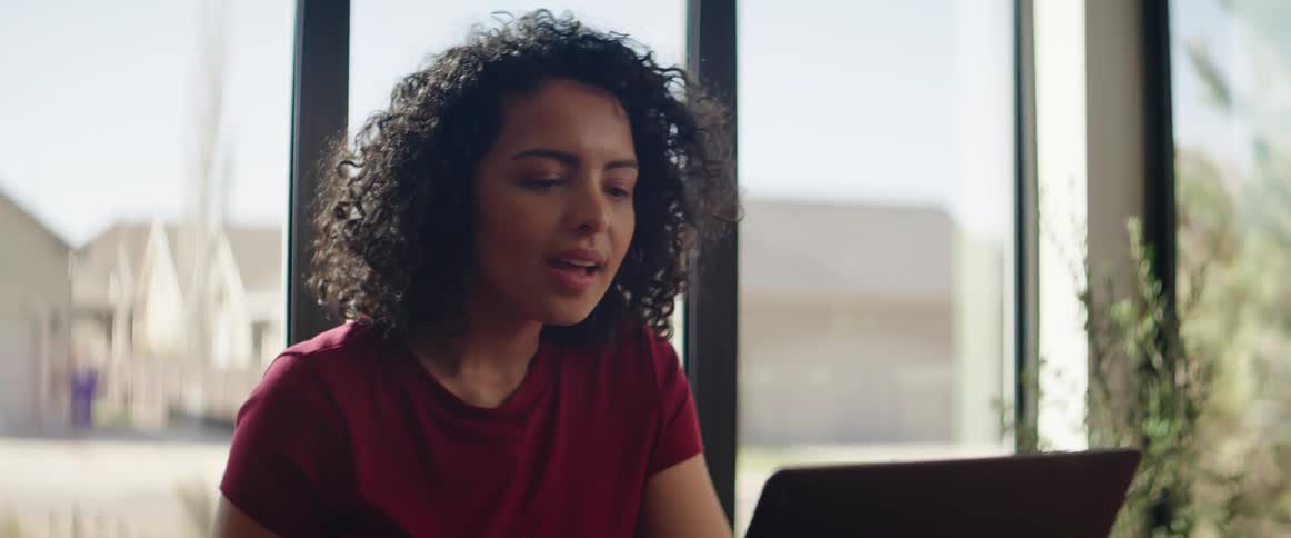 A black teenager sits down at a table and reads on her laptop