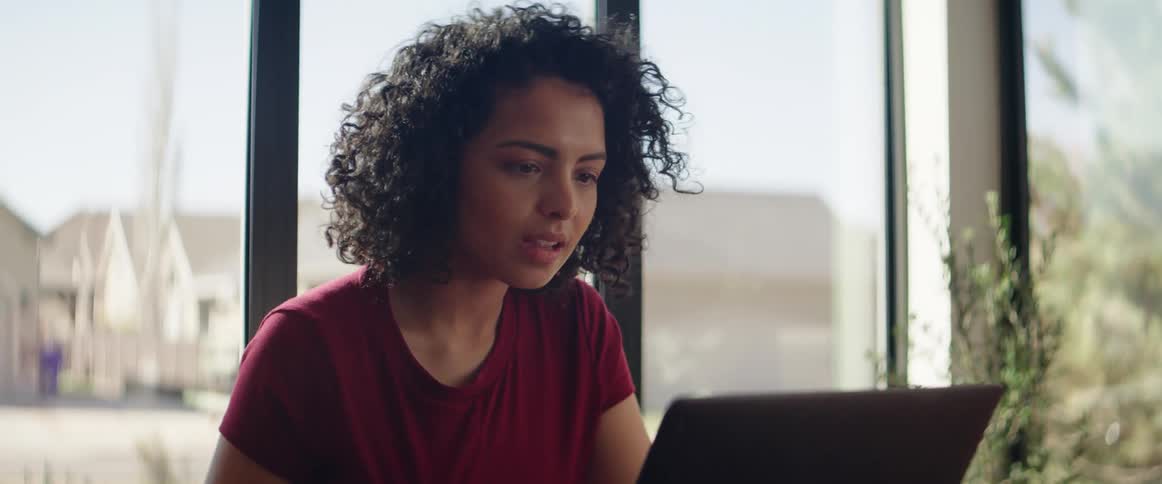 Female sits down at kitchen table then places laptop on the table opens it and starts to type and read the screen while smiling slightly