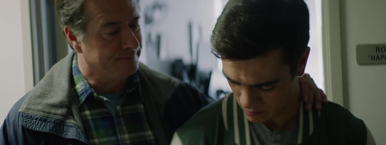 A man comforts his teenage son as they stand in the doorway of a hospital room