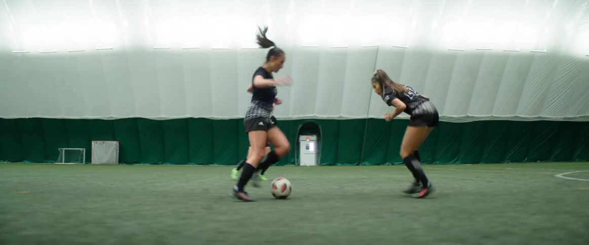 Three female soccer players practice running and kicking a ball in an indoor soccer field