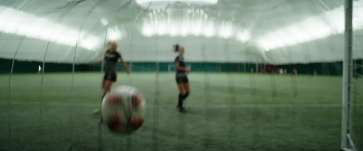 Two teenage soccer players practice kicking a soccer ball into a net in an indoor stadium