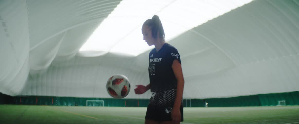 A female soccer player is practicing her footwork with a soccer ball while standing on the sidelines of an indoor soccer field
