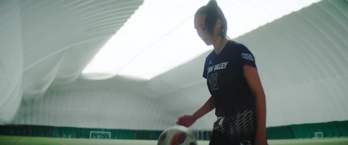 A female athlete repeatedly kicks a soccer ball in an indoor arena
