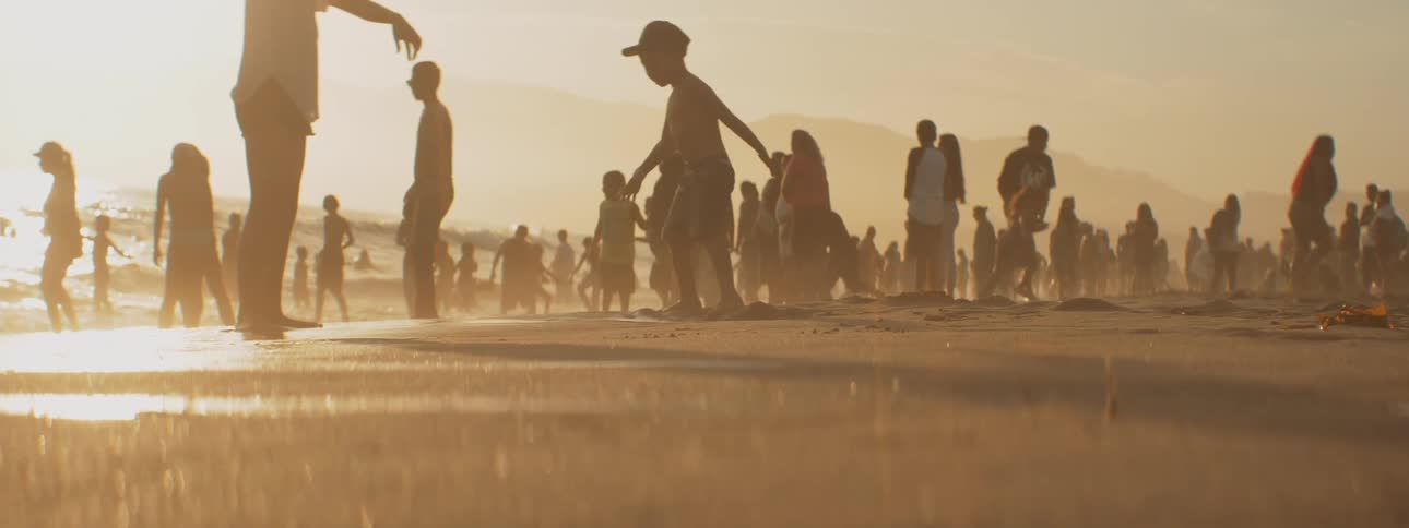 A crowd of adults and children play on the beach and swim in the ocean as the sun starts to set in the horizon