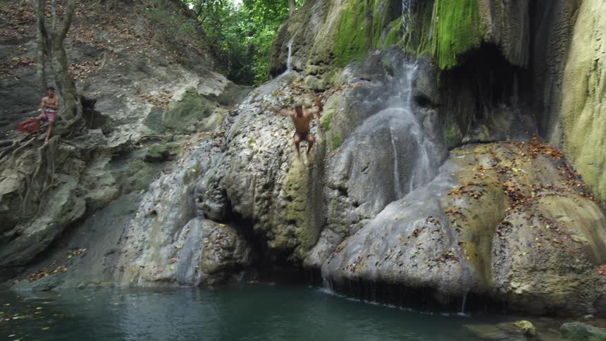 A white teen jumps off of rocks into a body of water as a friend watches