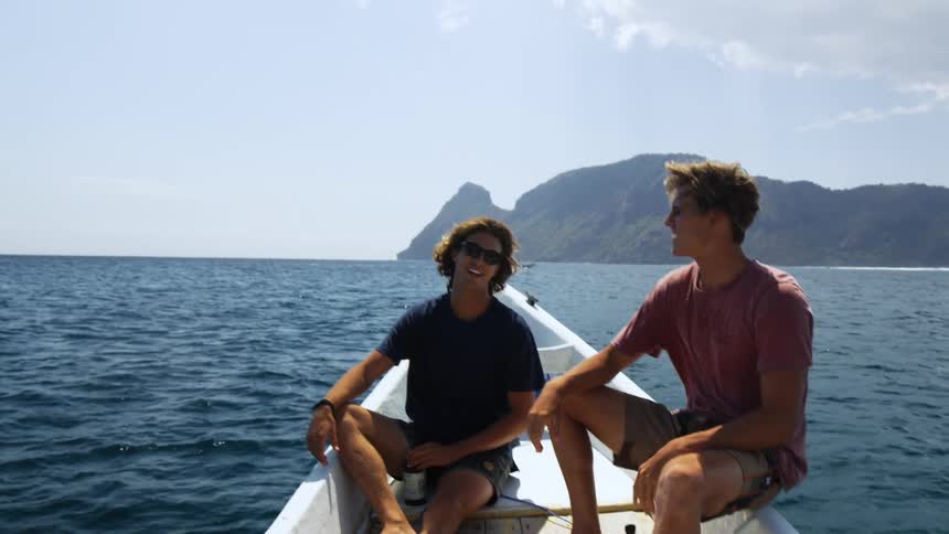 Two men sit in boat looking across ocean with mist hovering around shore in background