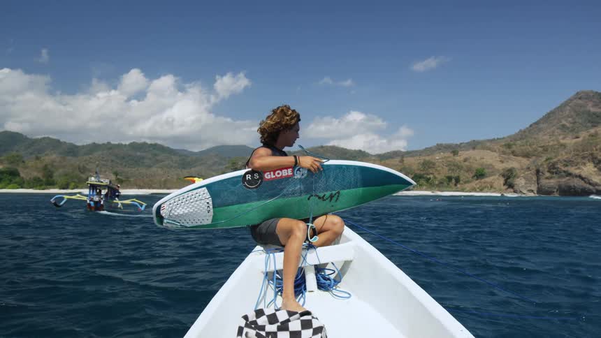 A man sitting on a boat holding a surfboard with land and sky in the background