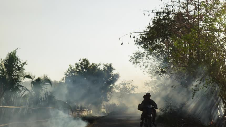 A motorcycle with passenger rides down a narrow road through billowing smoke from a brush fire
