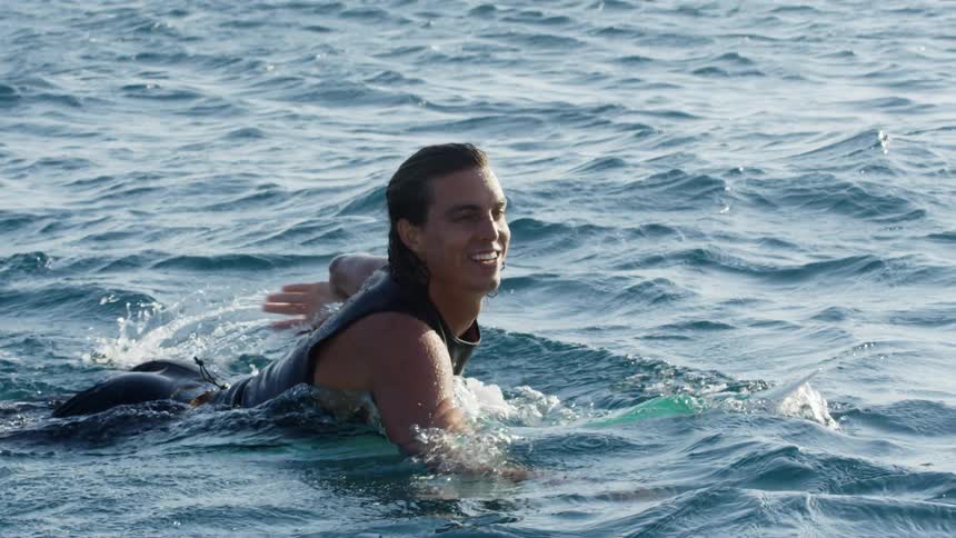 A young white male paddles through the ocean on a surfboard