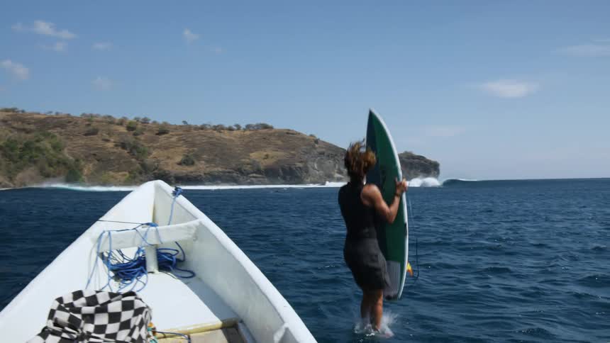 A man with a surfboard jumping off the boat into the ocean with land and the sky in the background