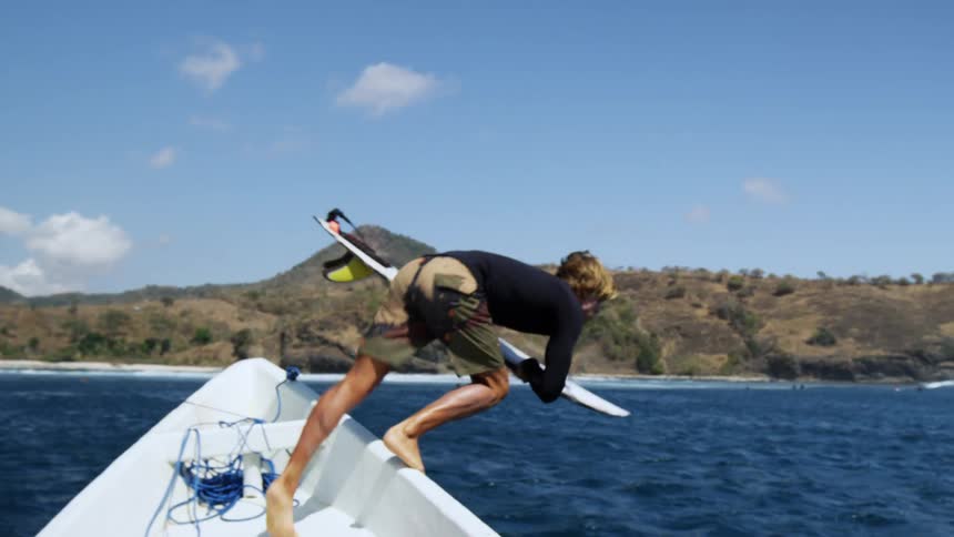 Man on a boat holds on to a surfboard and jumps into the ocean