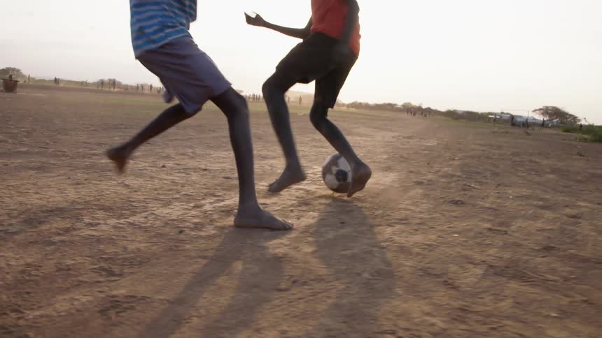 A group of children with short pants plays football barefoot on a dirt field