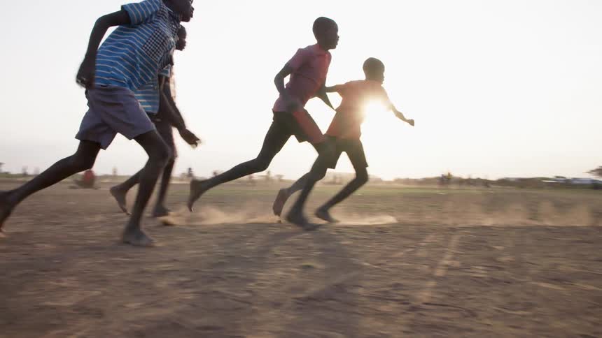 Several black boys play a game of soccer outdoors on a dirt plain