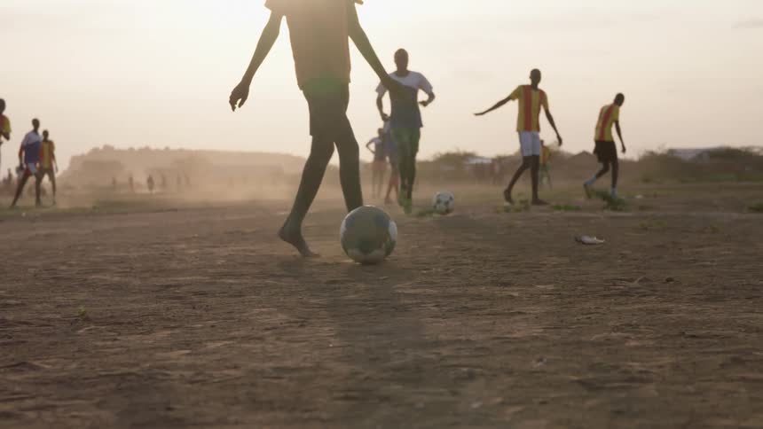 A number of black males play soccer in a dirt field