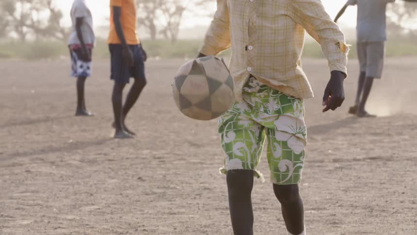 A barefoot boy dribbles a soccer ball in the dirt while others play behind him