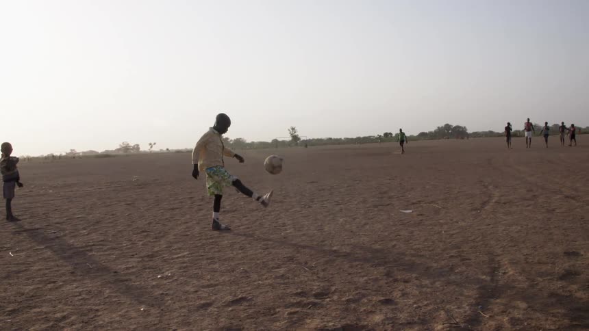 A young black boy kicks a soccer ball around on a dirt plain