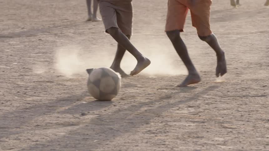 Barefoot boys kick around a tattered soccer ball in the dirt on a sunny day