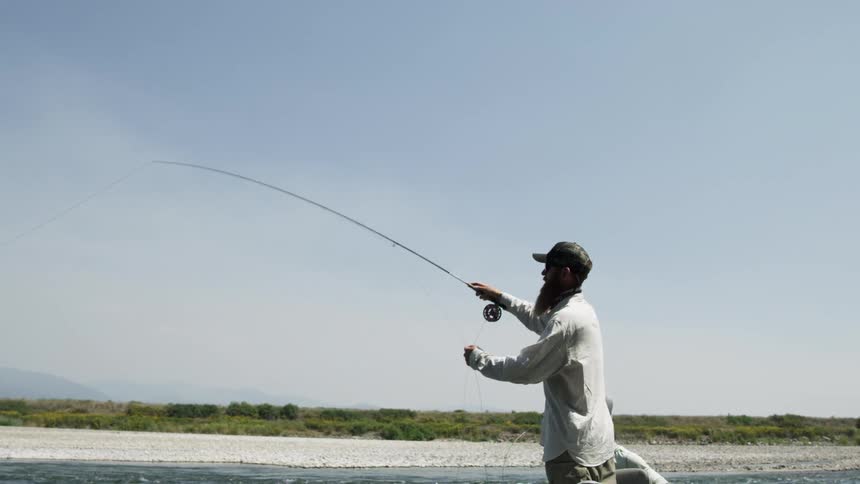 A white man stands in a boat and casts a fishing line into a lake