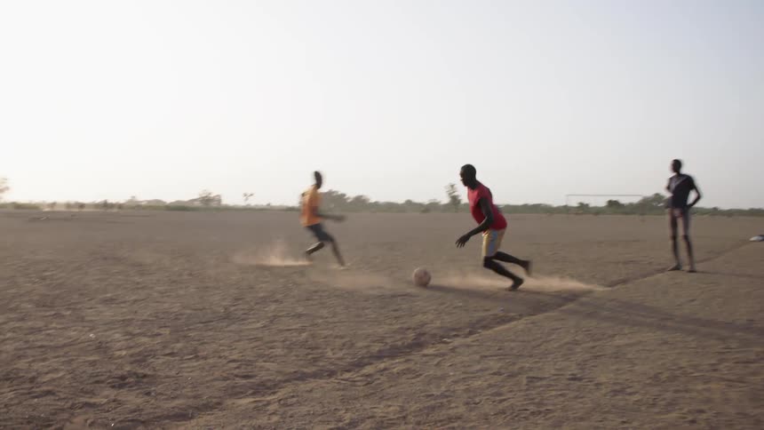 A group of black men in casual clothing playing soccer barefoot on dirt soccer plot