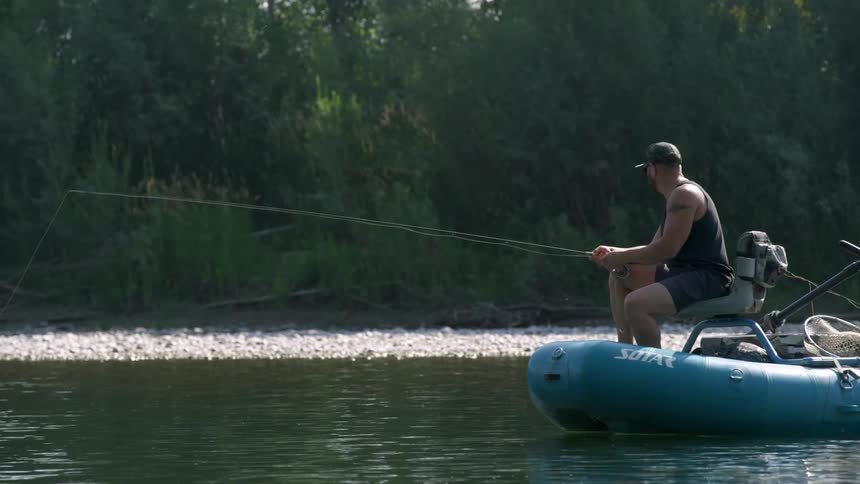 A man sits on a small boat in a lake holding a fishing pole out to the water