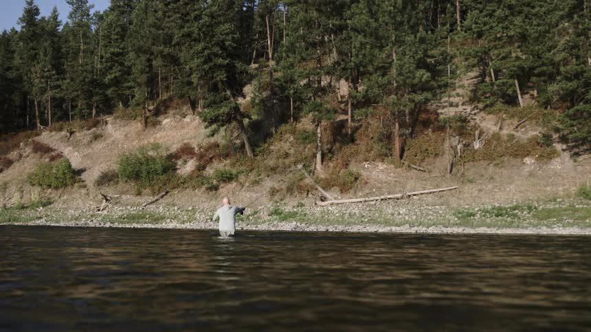 A white man stands waist deep in a river and fishes near a shoreline