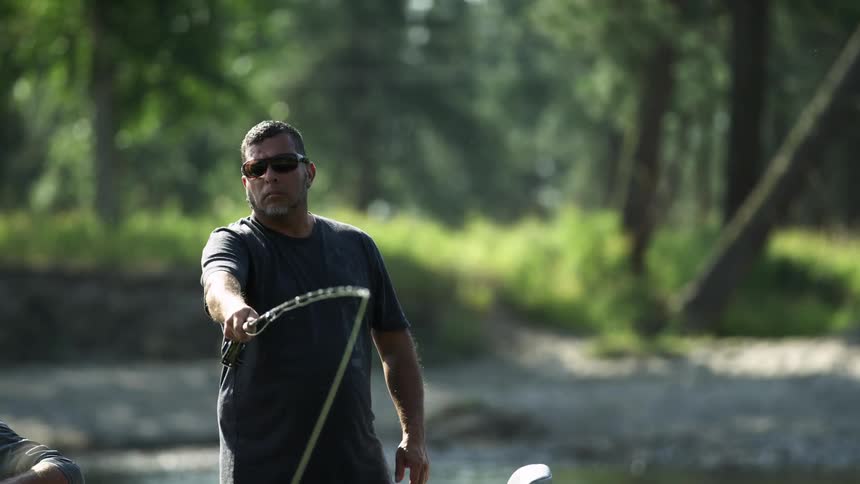 A man with sunglasses fly fishing with another man next to him with trees and plants in the background