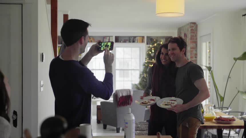 A man takes a photo of a couple holding up plates of food before they sit down to eat