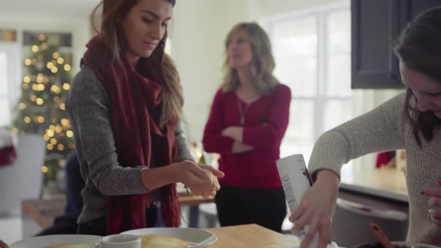 Two women prepare food while another woman and two men talk nearby as a christmas tree sits at the end of the room
