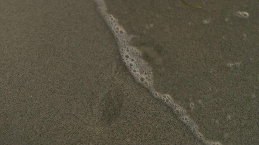 A small wave washes over a footprint in the sand