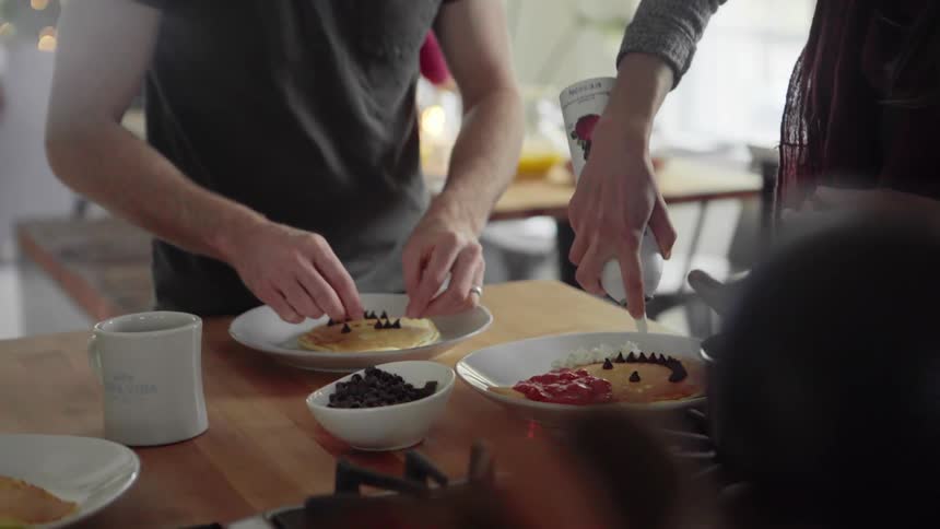 Two family members decorate pancakes with whipped cream and berries in a kitchen