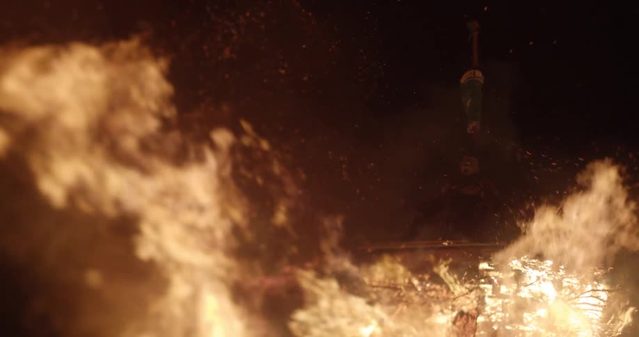 Fire burning tree branches in the foreground with a person sitting on a platform in the background