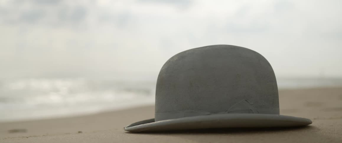 A derby hat sits on the wet beach sand as waves come to shore
