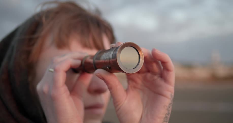 A woman looks through an old fashioned telescope while standing on shore