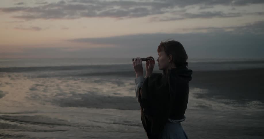 A white woman stands on a beach and raises a spyglass to her eye