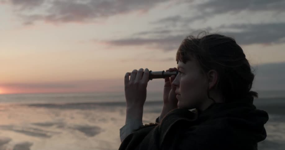 Woman standing on the wet sands of a beach uses a telescope to look at the horizon