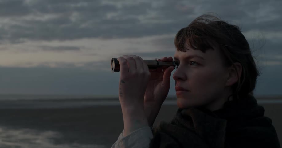 A woman with a telescope standing on the beach with the sky in the background