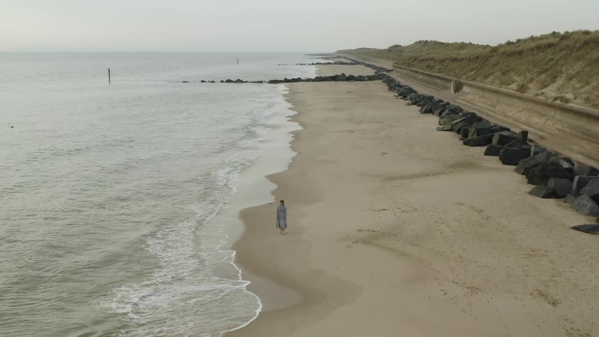 A woman in a light blue dress walks along a beach just out of reach of the waves
