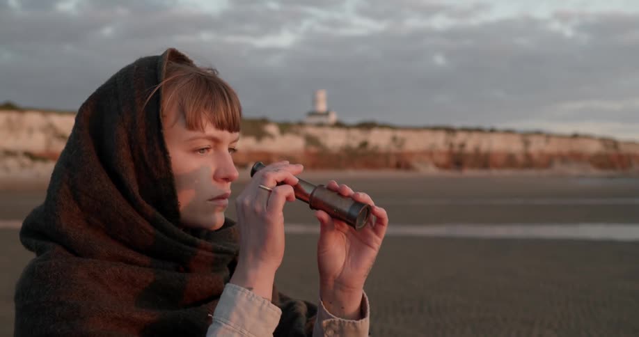 A woman on the beach looking through a telescope with the sky and clouds in the background