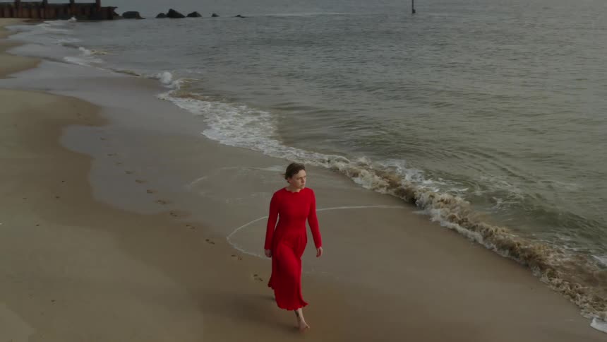 A white woman in a red dress walks barefoot on a beach