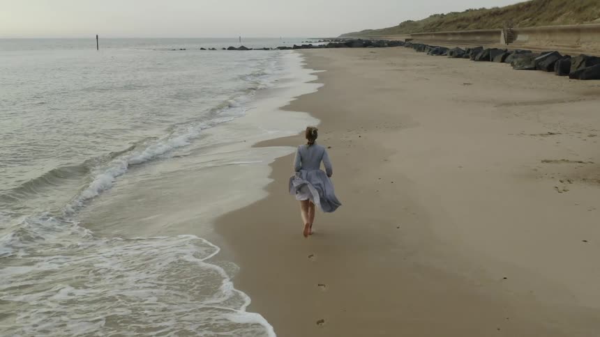 A woman holds up the front of her dress as she runs barefoot on the wet sand of the beach