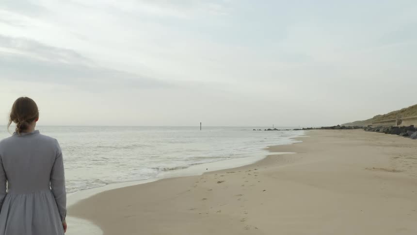A woman stands on the edge of the beach in a long blue dress as waves crash onto the shore