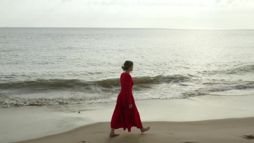A white woman in a red dress walks on a beach as the tide comes in