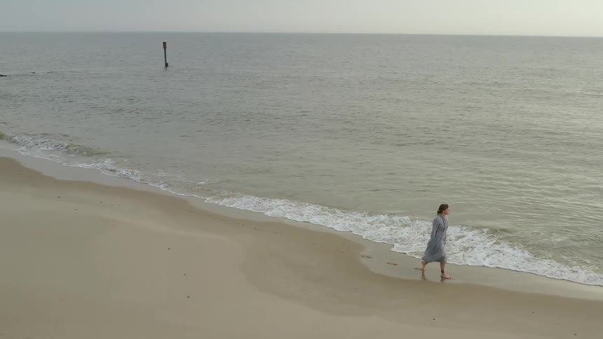 A white woman in a dress walks barefoot on a beach