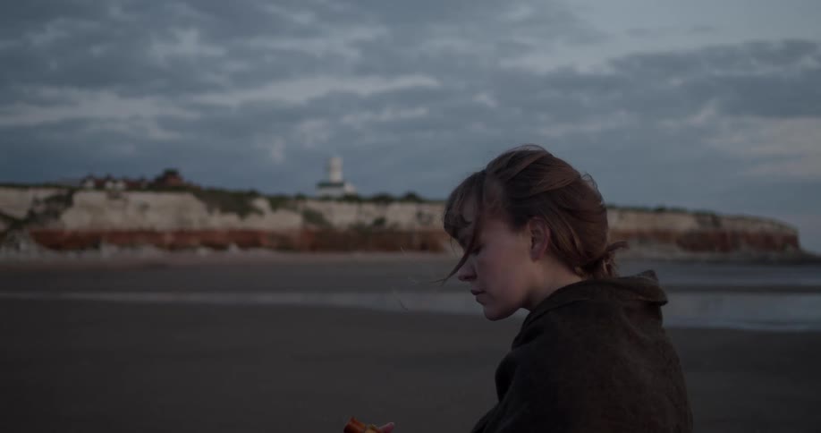 A woman walks on the beach on a cloudy day holding a bottle
