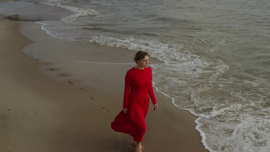 Woman wearing long red dress walks barefoot on the shoreline of a beach