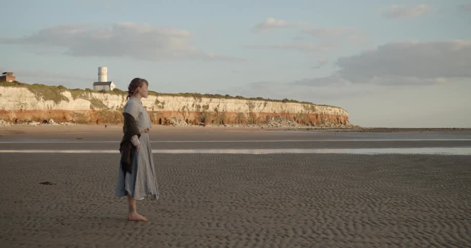 Woman stads barefoot on the sands of a beach near a cliff at sunset