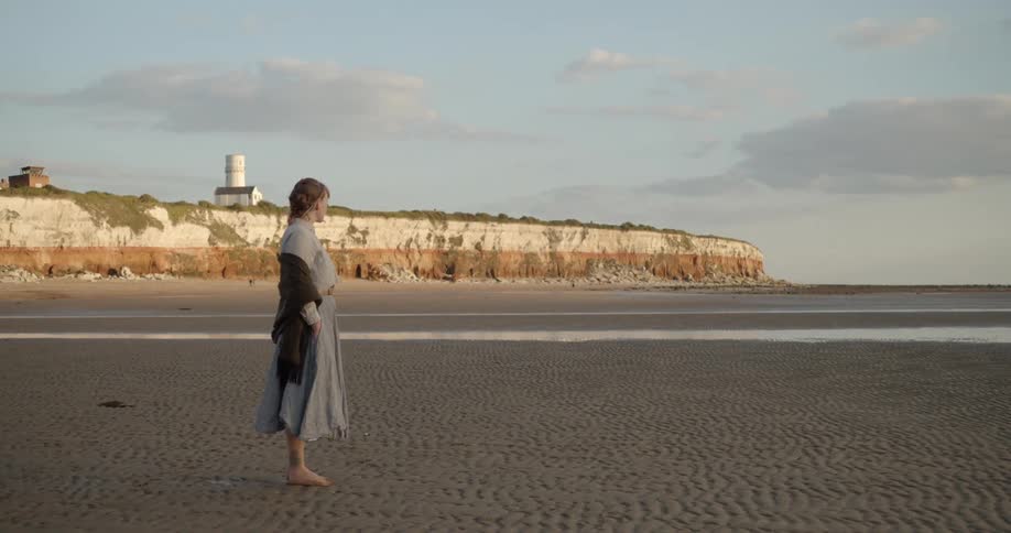 A woman walks on hard sand below a seaside cliff