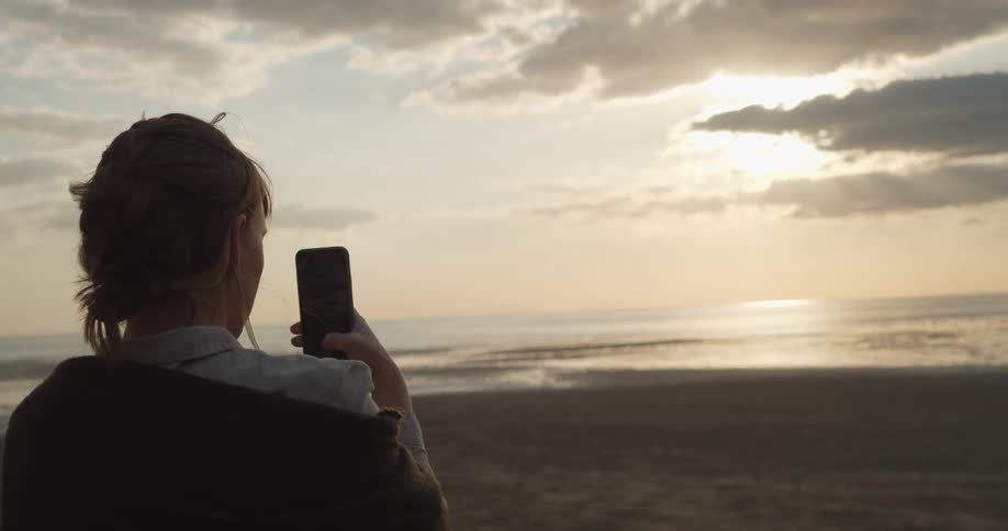 Woman standing on a beach takes a photo of the sunset on her phone