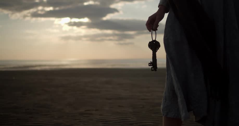 A woman holds a key chain of three large old fashioned keys while standing on a beach facing the ocean