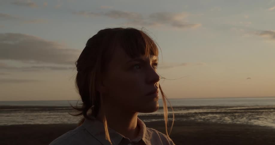 A woman stands on shore staring and thinking as a breeze blows her hair